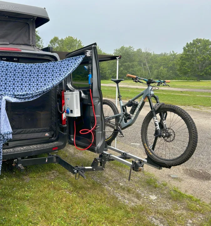 This RV rental camper van features a pop-top, outdoor shower, and a mountain bike on its rear rack, parked in a grassy area.. Ford Custom Transit 2022
