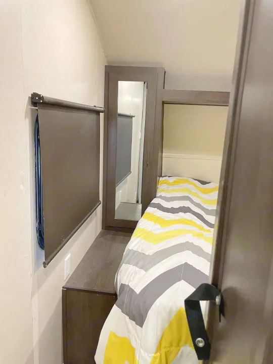 This RV rental bedroom offers a cozy bed with a yellow and gray striped comforter, dark window blind, and built-in storage.. Forest River Wildwood X-Lite 2019