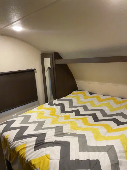 An RV rental bedroom boasts a bed with a yellow & gray chevron comforter, practical window blind, and built-in storage.. Forest River Wildwood X-Lite 2019