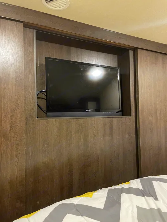RV rental bedroom: TV and soundbar integrated into wood cabinetry with sliding panels. Bed with chevron duvet.. Forest River Wildwood X-Lite 2019