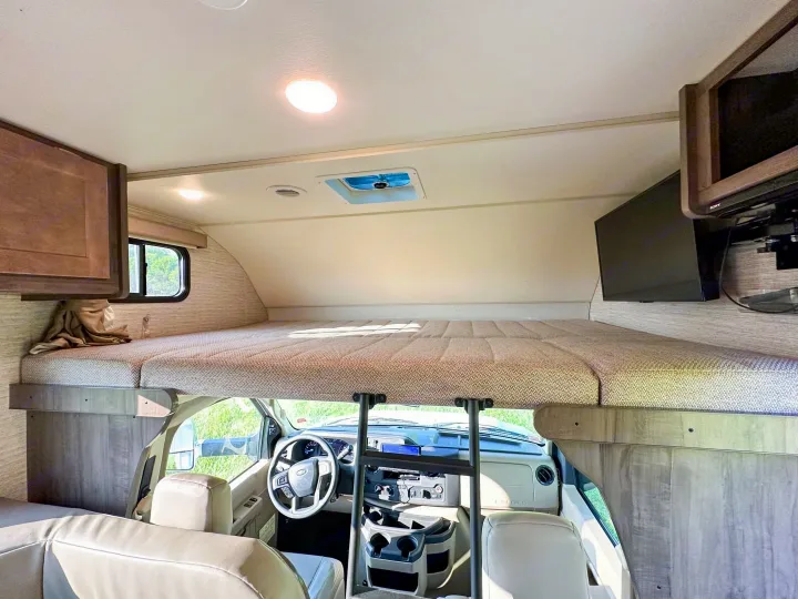 Inside an RV rental: Looking forward to a cozy overhead bunk with patterned mattress, twin mounted TVs, and the driver's cab.. Winnebago Minnie Winnie 2021