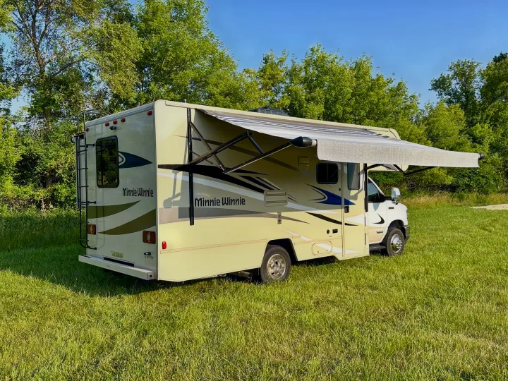Beige Minnie Winnie Class C RV rental with extended awning parked on a sunny grassy field.. Winnebago Minnie Winnie 2021