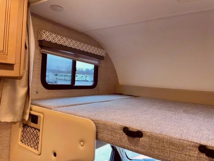 Inside an RV rental: a cozy over-cab bunk bed with a window and storage, ready for your next adventure.. Thor Coleman 2022