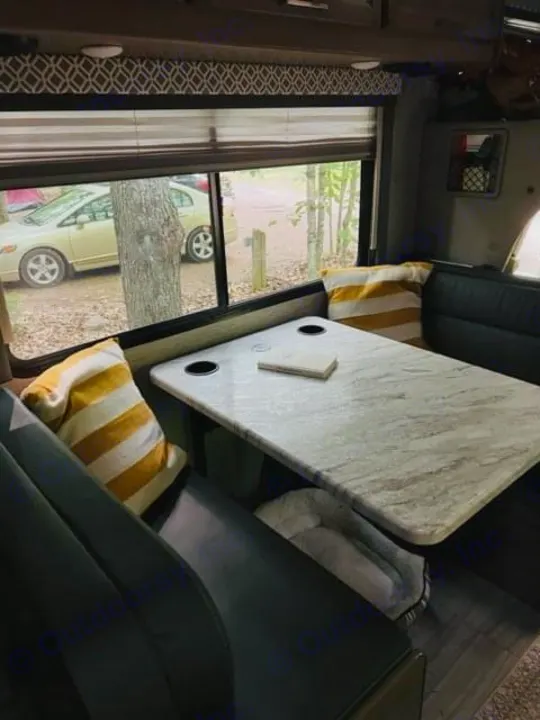 Experience RV rental comfort: a cozy dinette with a durable table, benches, and striped pillows, overlooking a campsite.. Thor Coleman 2022