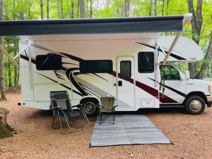 RV rental parked in a wooded campsite with awning, chairs, and mat, perfect for outdoor relaxation.. Thor Coleman 2022