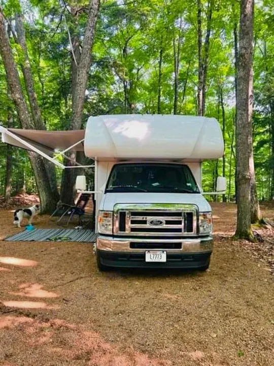 A white RV rental, a Class C motorhome, is parked in a treed campsite with its awning extended and a small dog resting nearby.. Thor Coleman 2022
