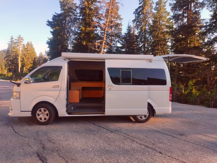 White campervan, ideal RV rental, parked among tall trees. Side door open reveals wood interior; rear hatch also open for outdoor adventures.. Toyota Hiace 2005