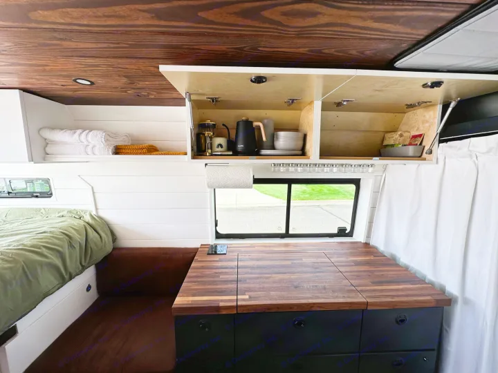 This RV rental showcases a cozy van interior with a bed, shiplap walls, wood ceiling, and a functional kitchen with opened cabinets.. RAM ProMaster 2500 2022