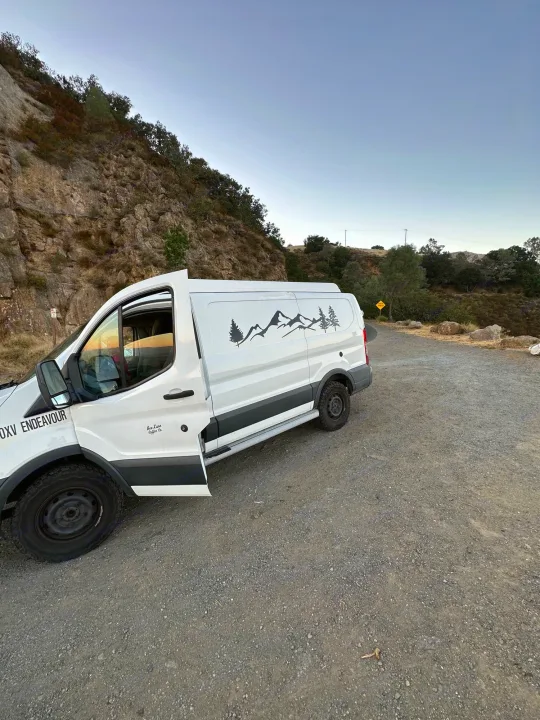 White camper van (RV rental) with mountain decals parked on a gravel road beside a rocky hillside.. Ford Transit 2016
