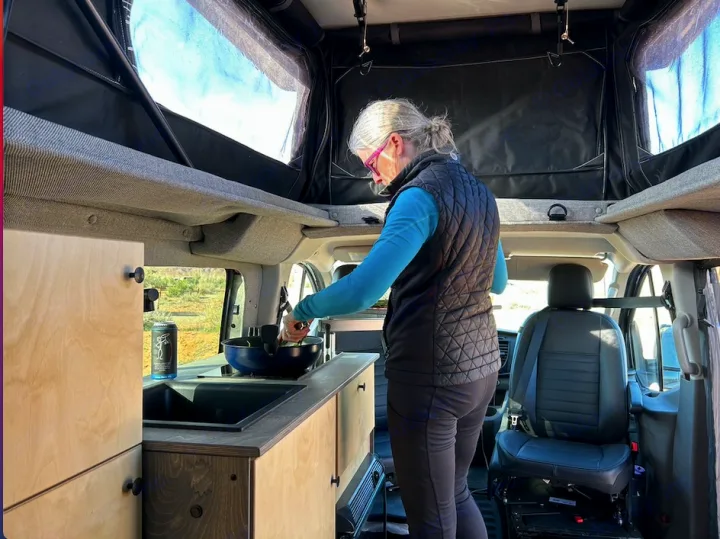 A woman cooks in a spacious RV rental van with a pop-top roof, ideal for comfortable van life adventures.. Ford Custom Transit 2022
