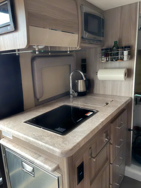 Bright RV kitchen with light wood-grain cabinets, induction cooktop, sink, microwave, spice rack, and fridge. Perfect for an rv rental.. Thor Motor Coach Scope 18M 2023