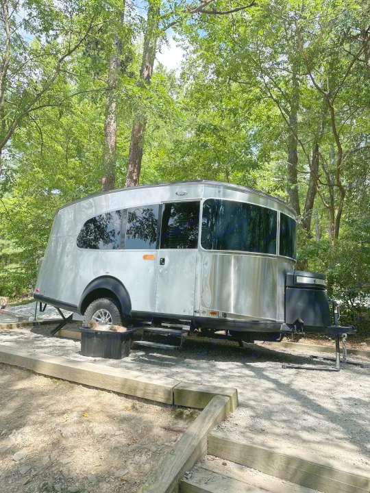 Silver Airstream RV rental parked on a sunny gravel campsite, surrounded by lush green trees. Enjoy nature camping.. Airstream Basecamp 2022