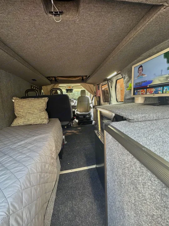 Inside this rv rental, a comfortable van conversion features a bed, entertainment monitor, and carpeted walls.. Ford Transit 2016