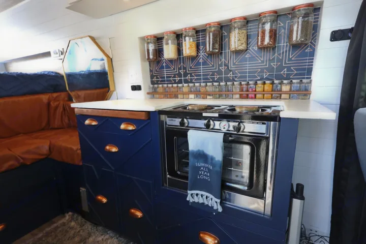 Stylish RV rental with a compact navy kitchen, built-in oven, spice rack, and a cozy brown leather lounge area. Van life ready.. Ford Transit 2019