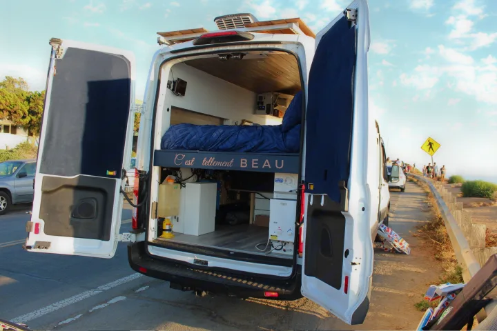 Open rear doors of a customized white camper van, showcasing its cozy bed, wood interior, and utilities, perfect for an rv rental.. Ford Transit 2019