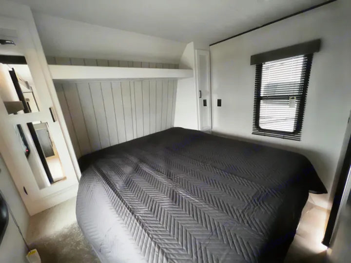Cozy RV rental bedroom featuring a large bed with black quilted comforter, modern white interior, and window.. Keystone Hideout 2022