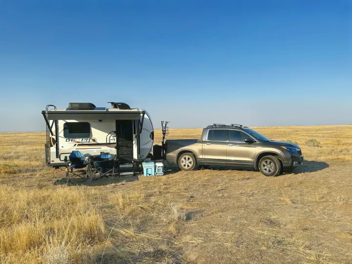 A tan truck next to a white GEOPRO rv rental, parked with camping gear in a vast, dry grassy field under clear skies.. Forest River Rockwood Geo Pro 2023