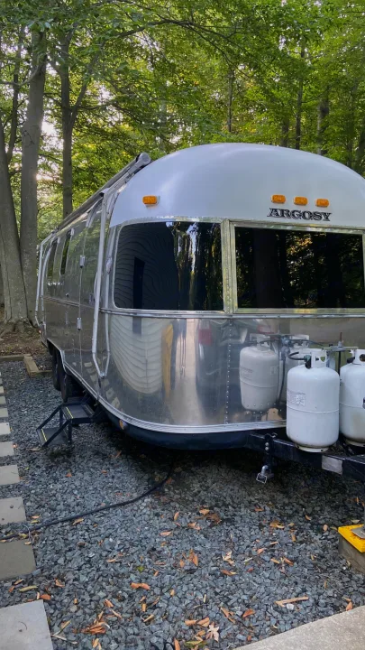 Silver & white Airstream Argosy RV rental with propane tanks, nestled on gravel in a peaceful, wooded setting.. Airstream Argosy 1977