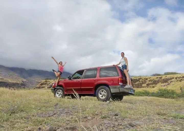 Two women playfully pose with a red SUV on a grassy hill, mountains behind. Off-road adventure awaits, no RV rental needed for this fun!. Xplorer Explorer 2007