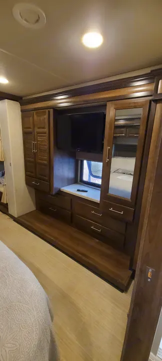 This RV rental bedroom features a mounted TV, storage cabinets, and drawers. A comfortable bed is in the foreground.. Entegra Coach Vision XL 2020