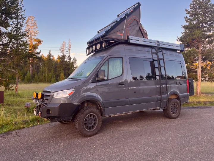 Gray adventure van RV rental with a rooftop tent, solar panels, and off-road tires, parked by pine trees.. Mercedes-Benz Sprinter 2019