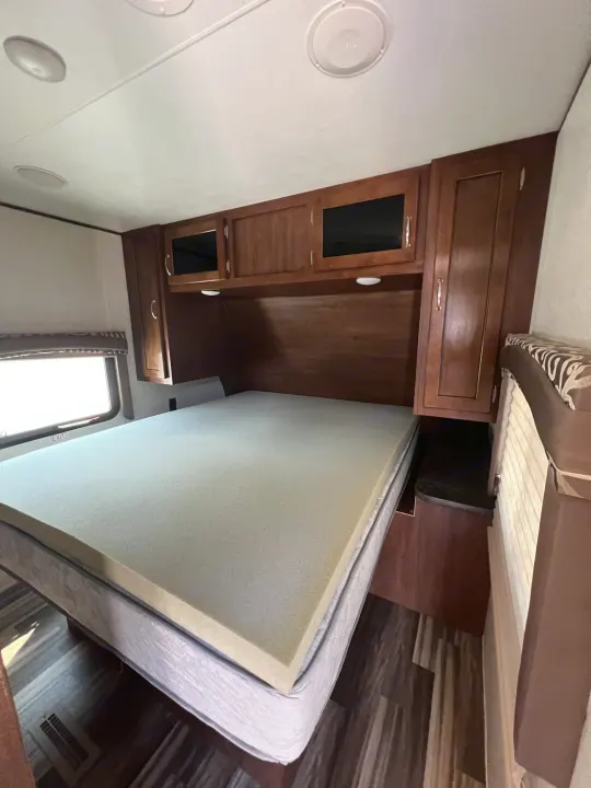 Inside an RV rental: a cozy bedroom with a bed, foam topper, overhead wood cabinets, and window.. Jayco Jay Flight 2017