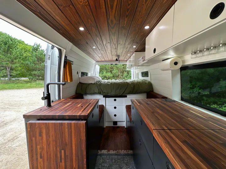 Bright RV rental interior features dark wood ceiling & counters, modern kitchen, & cozy rear bed.. RAM ProMaster 2500 2022