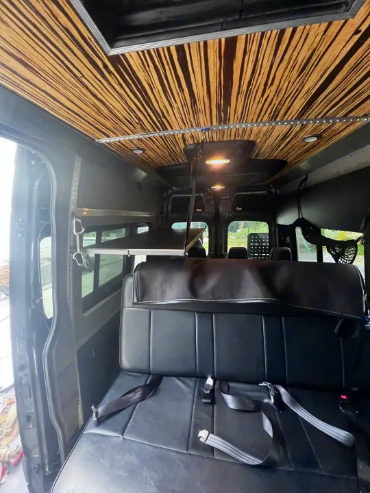 Inside an RV rental: a black bench seat and striped wood ceiling with LED lights. A fold-out bunk/shelf is on the left.. Mercedes-Benz Sprinter 2019