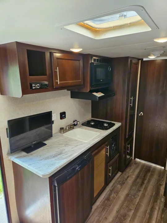 Well-equipped RV rental kitchen featuring dark wood cabinets, light counters, sink, stove, microwave, and TV.. Jayco Jay Flight SLX 2019
