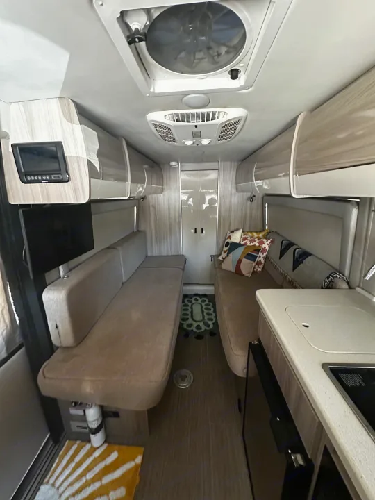 Clean RV rental interior: parallel sofas, compact kitchen on right, monitor & TV on left, overhead fan & AC.. Thor Sequnce 2023