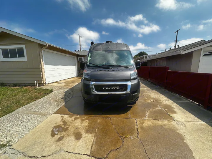 Front view of a dark grey Ram ProMaster van, ideal for an RV rental, parked in a sunny residential driveway.. Thor Sequnce 2023