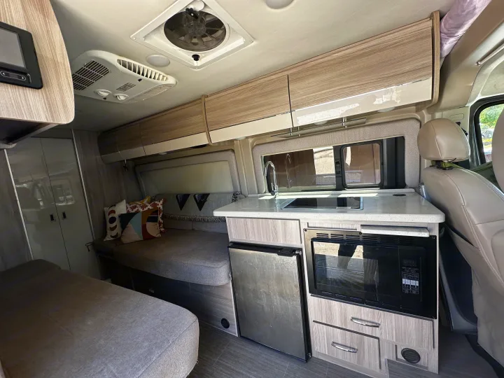 Inside an RV rental, showing a functional kitchen with appliances, comfortable seating/bed area, and overhead storage.. Thor Sequnce 2023