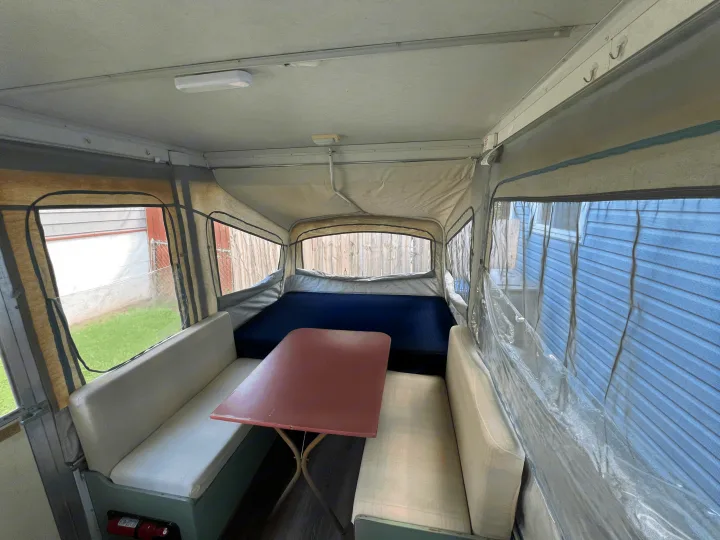 Inside an RV rental pop-up camper, showing a compact dining area with a red table and benches, and a blue bed.. Coleman Destiny Cedar 1992