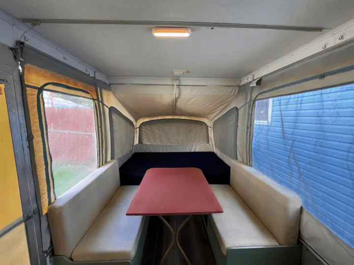 Interior of a pop-up RV rental: dining table, benches, and bed within a canvas-walled camper with clear windows.. Coleman Destiny Cedar 1992