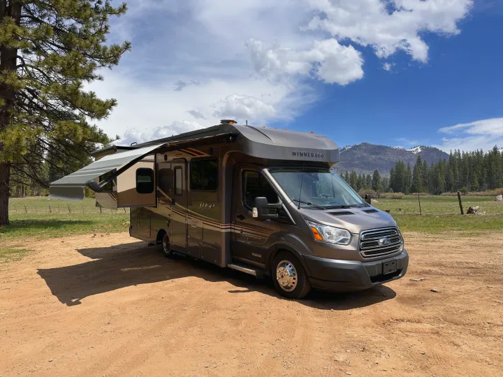 Winnebago Fuse rv rental parked on a dirt road with its awning extended, surrounded by trees, mountains, and blue sky.. Winnebago Fuse 2017