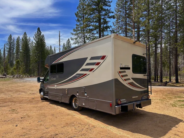 A gray and beige RV rental, a Winnebago Fuse, is parked on a dirt road in a pine forest under a blue sky.. Winnebago Fuse 2017