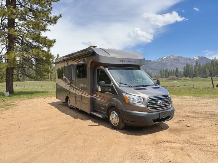 This gray Winnebago Fuse Class C rv rental sits on a dirt road, framed by pine trees, a field, and mountains.. Winnebago Fuse 2017