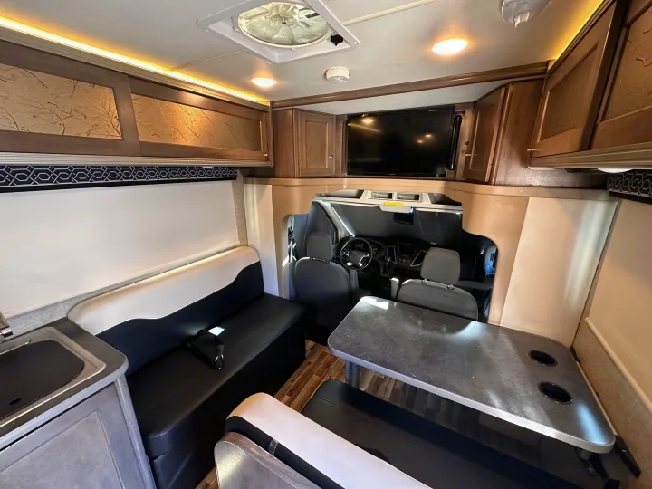 Bright rv rental interior featuring a cozy dining area, kitchenette, overhead storage, and a glimpse of the driver's cabin.. Winnebago Fuse 2017