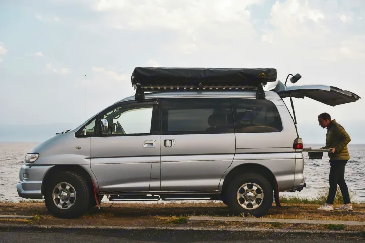 Silver campervan with roof awning parked seaside. Person cooks at open rear hatch. Ideal for RV rental adventures.. Mitsubishi Delica 2005