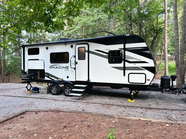 White Imagine XLS RV rental travel trailer parked at a wooded campsite, featuring outdoor kitchen.. Grand Design Imagine XLS 2023