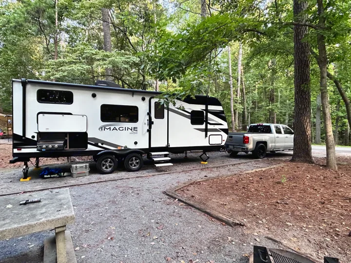 Campsite ready: A white RV rental, truck, and outdoor kitchen grill set up in a lush forest.. Grand Design Imagine XLS 2023