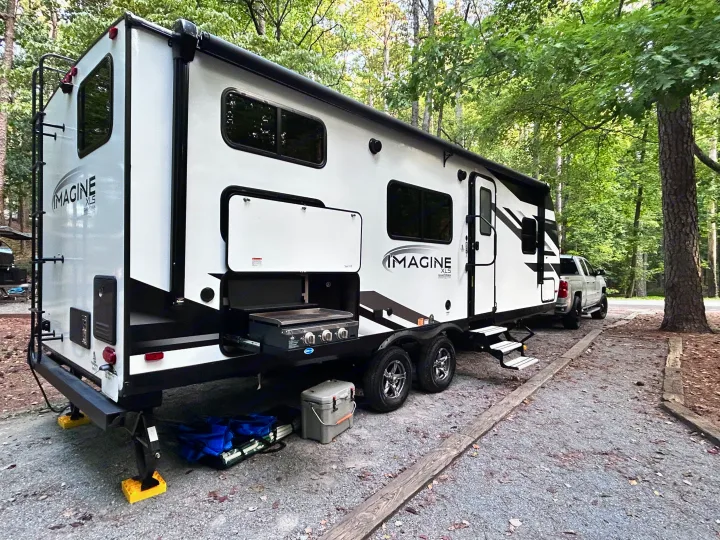 Imagine XLS RV rental parked at a wooded campsite, featuring an outdoor grill, ready for camping adventures.. Grand Design Imagine XLS 2023