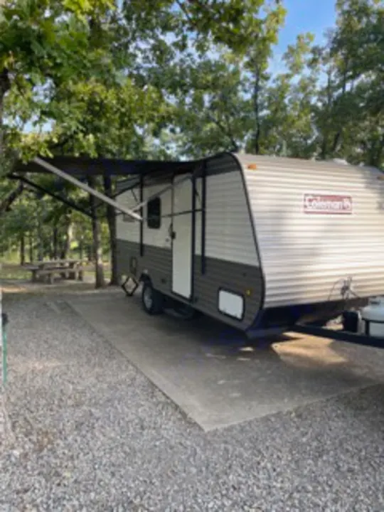 A Coleman RV rental is parked at a wooded campsite with its awning extended, perfect for outdoor travel.. Dutchmen Coleman Lantern LT 2023