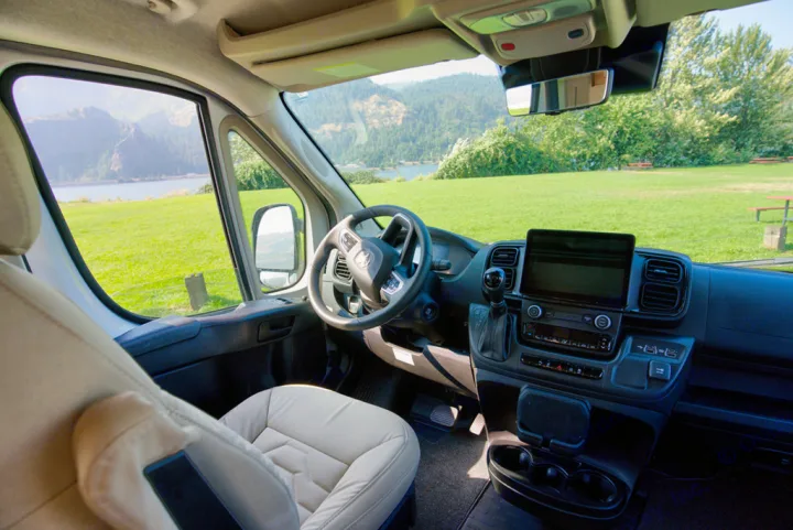 RV rental's spacious cockpit: beige seats, dash, and a beautiful view of a lake, mountains, and green landscape.. Coachman Nova 20RB 2023