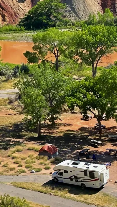 Overhead view of a bustling campground: an RV rental, tent, and picnic tables nestled amidst trees, with a muddy river and mountains.. Jayco Redhawk 2017