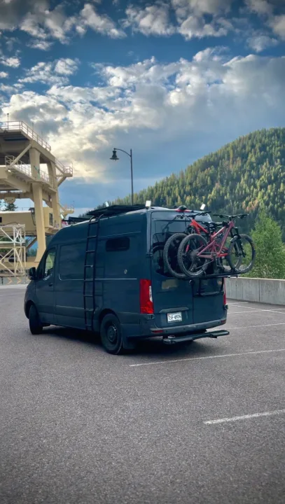 Dark blue Mercedes Sprinter camper van with mountain bikes parked near a large industrial building and forested mountains. An ideal rv rental.. Mercedes-Benz Sprinter RV Motorhome Campervan 2019