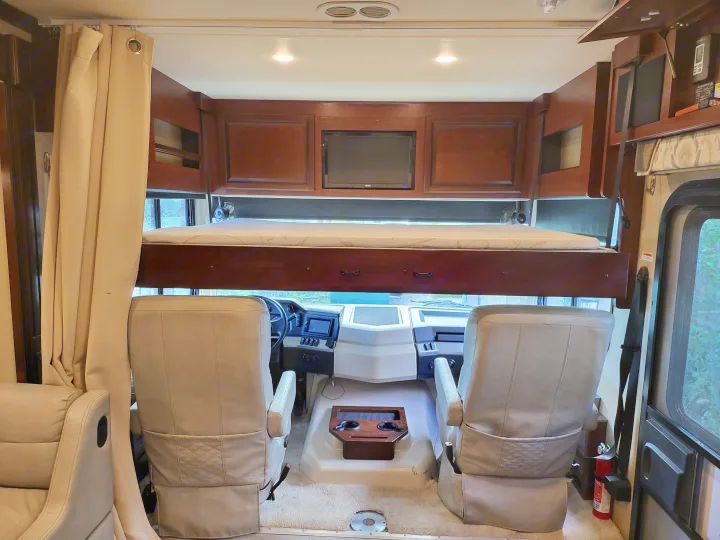 Inside an RV rental: front cockpit area with cream seats, an overhead bunk bed, and wooden cabinets, ideal for travel.. Fleetwood Bounder 2017