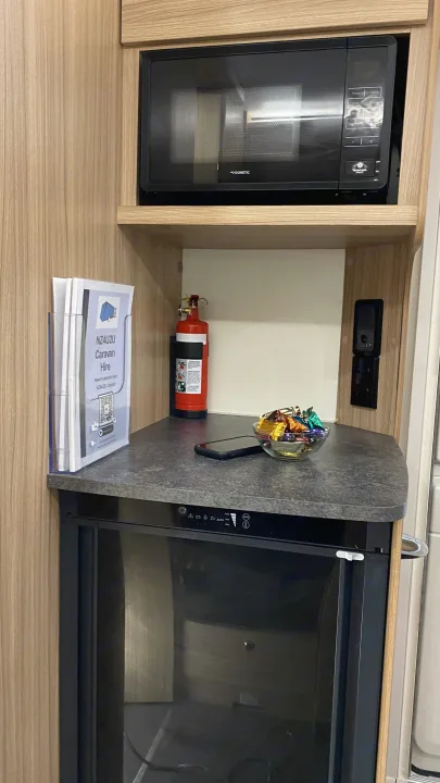 Compact kitchen setup in an rv rental, featuring a Dometic microwave, refrigerator, fire extinguisher, hire brochures, and a bowl of candies.. Bailey Pheonix 640 2022