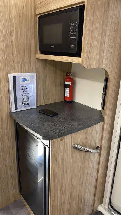 Inside an RV rental, a compact kitchen features a Dometic microwave, fridge, fire extinguisher, and NZ4U2U caravan instructions.. Bailey Pheonix 640 2022