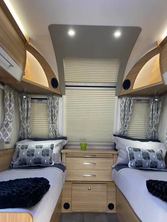 This bright RV rental bedroom features twin beds, a wood nightstand, patterned curtains, and ambient lighting in the overhead cabinets.. Bailey x 2022